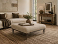 Upholstered square ottoman coffee table with a textured fabric finish and turned wood legs, styled in a cozy transitional living room.