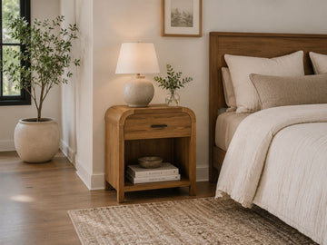 Natural wood one-drawer accent table styled as a bedside table with a ceramic lamp in a modern bedroom.