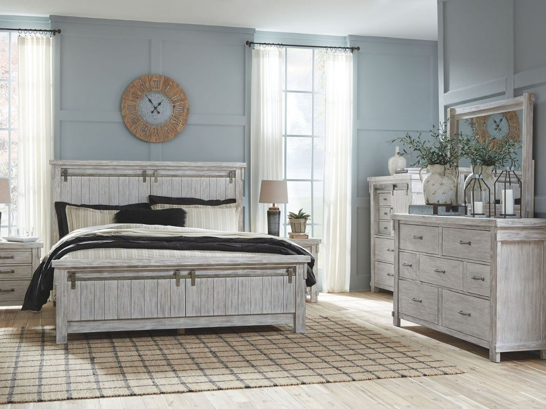 A rustic, white-washed wooden panel bed featuring a tall headboard and matching footboard with vertical planking and decorative sliding barn door hardware.