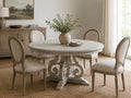 Round whitewashed wood dining table with an ornate pedestal base and four upholstered dining chairs.