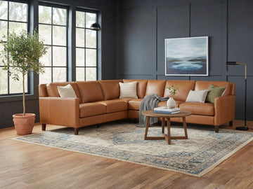 A large, L-shaped corner sectional in a warm cognac leather finish, styled with a round wooden coffee table and a tall olive tree.