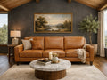 A three-cushion cognac leather sofa is centered against a dark charcoal-gray accent wall. A large, gold-framed landscape painting hangs above the sofa, and a round marble coffee table with a dark wood base sits in the foreground.
