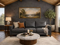 A three-cushion charcoal gray leather sofa is set against a dark, textured charcoal accent wall. A gold-framed landscape painting of a river hangs above the sofa, illuminated by warm light from a side lamp.