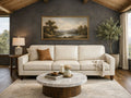 A bright cream leather three-cushion sofa stands against a dark charcoal-gray accent wall. Above the sofa hangs a large, horizontally oriented landscape painting of a river scene framed in gold. In front sits a round marble-topped coffee table with a dark wood base on a light neutral rug.