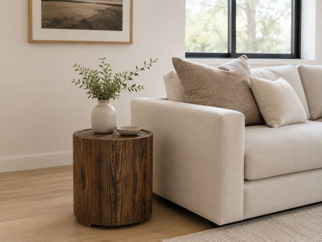 Dark brown round wood end table with vertical slatted detailing, styled beside a cream sofa in a warm contemporary living room.