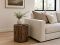 Dark brown round wood end table with vertical slatted detailing, styled beside a cream sofa in a warm contemporary living room.