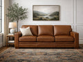 Brown leather three-seat sofa with plush cushions, set in a bright living room with light walls.