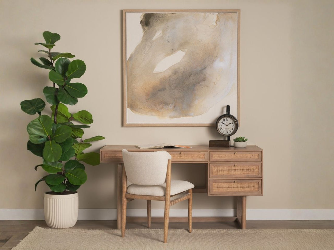 A styled home office scene featuring a light wood desk with cane-front drawers and a matching upholstered chair.