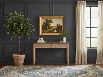A light, sand-toned wooden console table with two drawers and slender, tapered legs, positioned in front of a dark grey wall with traditional molding.