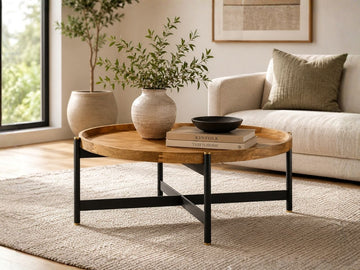 Round tray-top coffee table featuring a natural wood finish and black metal base, styled in a contemporary living room with neutral décor.