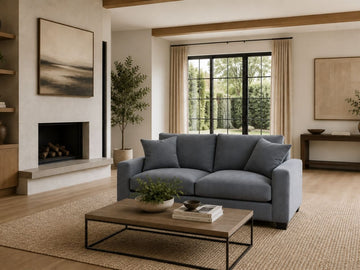 Blue upholstered sofa in a bright modern living room with a fireplace, large windows, wood accents, and a woven area rug