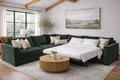 Green sectional is accented with neutral beige and white patterned throw pillows, maintaining the room's airy feel against the landscape painting and wooden beams.