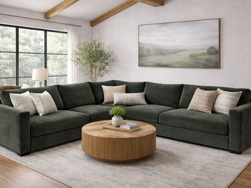 Large L-shaped sectional sofa in deep emerald green velvet fabric, styled with several neutral-toned decorative pillows. It is centered in a bright living room with vaulted wood-beamed ceilings and a large landscape painting on the back wall.