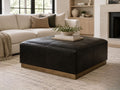 Black leather square ottoman with stitched detailing and a warm wood base, styled in a modern neutral living room.