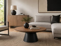 Modern round coffee table with a warm wood tabletop and sculptural black pedestal base, styled in a contemporary living room with neutral furnishings.