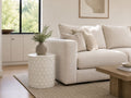 White cylindrical side table with a subtle geometric pattern, styled beside a cream sofa in a bright, modern living room.