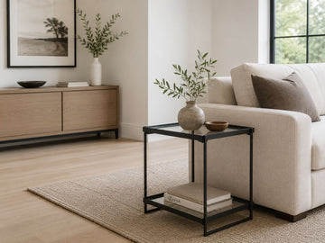 Modern black metal and glass accent table with a lower shelf, styled beside a cream sofa in a bright contemporary living room.