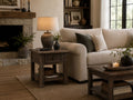 Rustic wood end table with a drawer and open shelf, placed beside a cream sofa in a warm farmhouse-inspired living room.