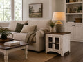 Rustic whitewashed farmhouse end table with cabinet storage, styled beside a cream sofa with a table lamp in a cozy living room.